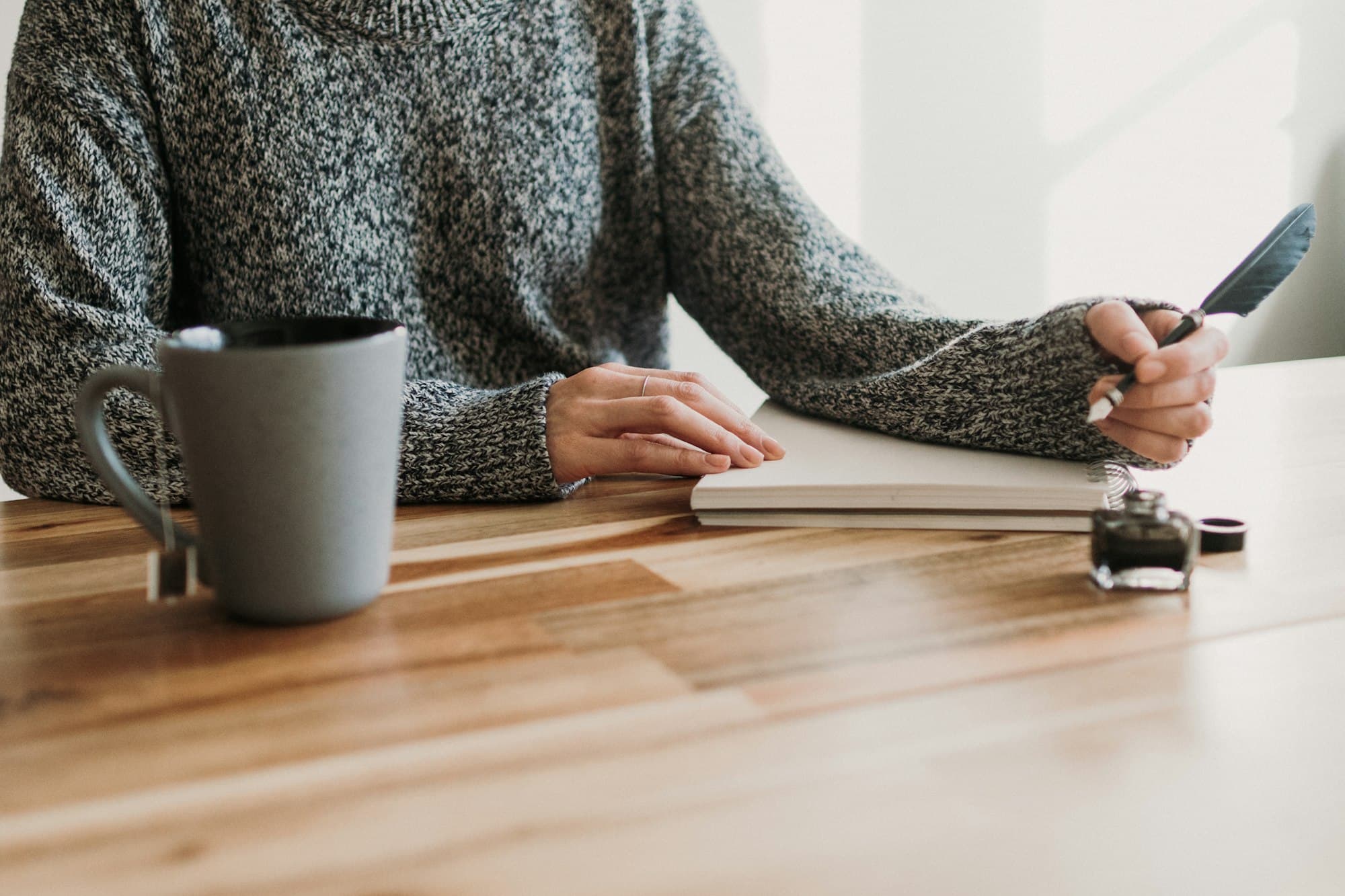 A person in a grey sweater writing at a light wooden table beside a grey mug and an ink bottle.