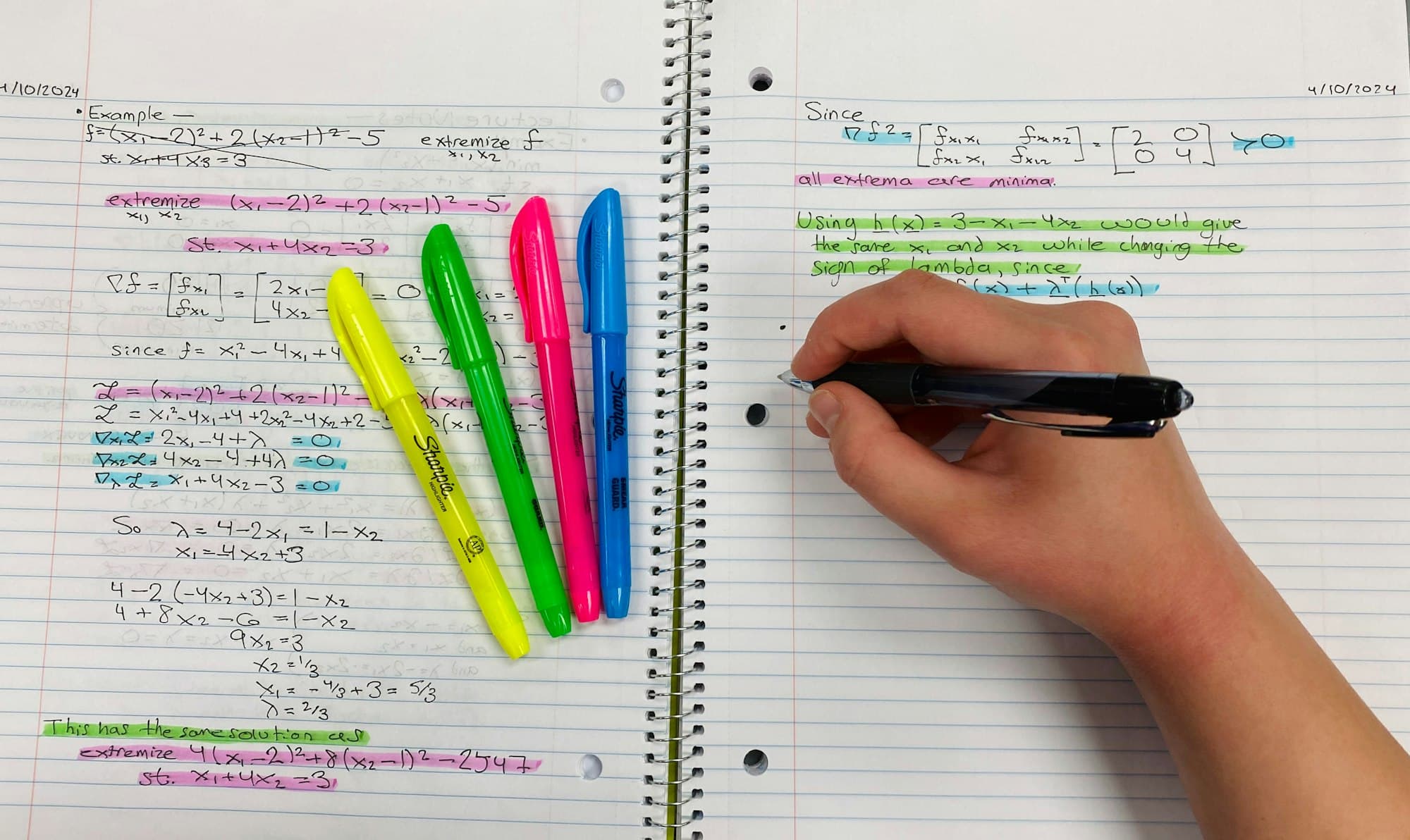 A hand writing in a spiral notebook full of worked notes, with yellow, green, pink and blue highlighters on the page.