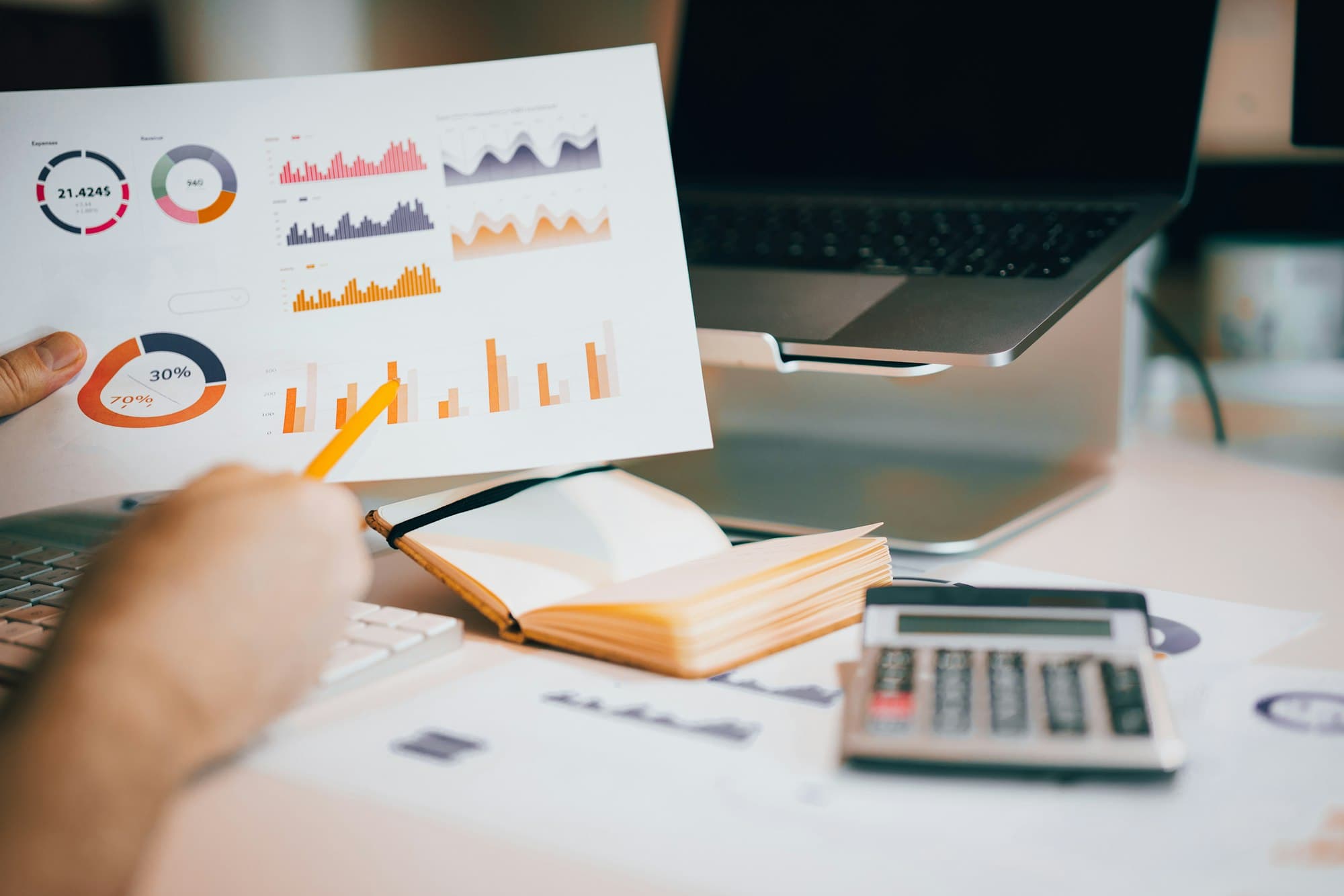 A hand holding a printed page of colorful charts above a desk with a calculator and laptop.
