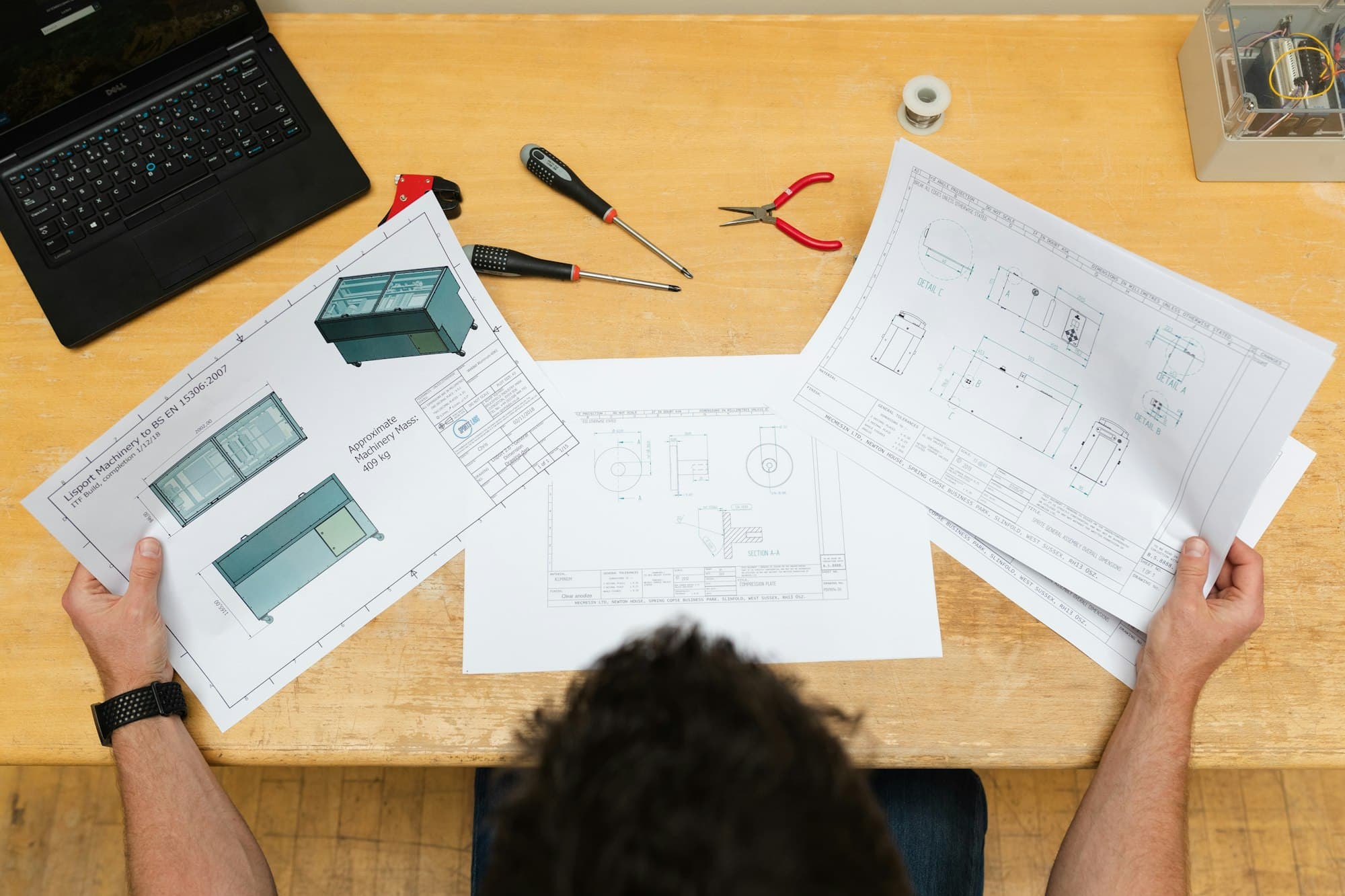 Seen from above, an engineer holds printed technical drawings at a wooden desk with screwdrivers and pliers.