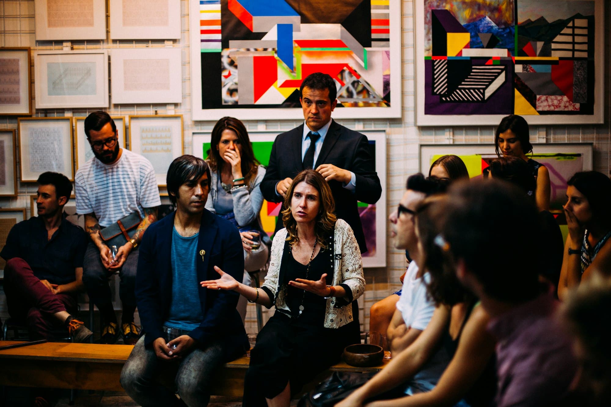 A group discussion in an art gallery, with people seated and standing before colorful geometric paintings.