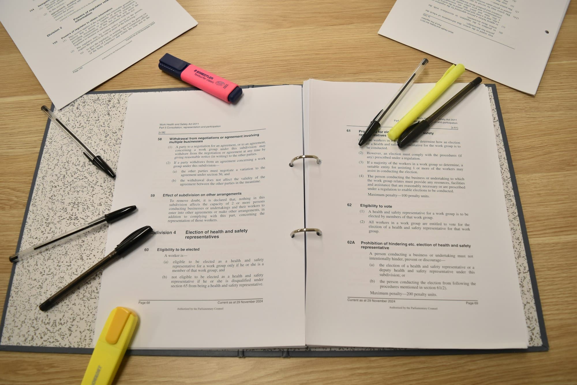 An open ring binder of printed pages surrounded by pens and pink and yellow highlighters on a wooden desk.