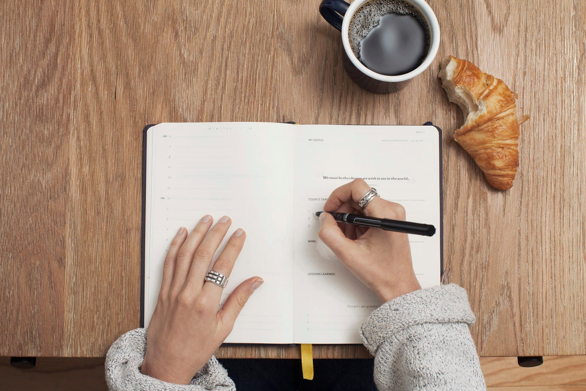 Seen from above, a hand writing in a notebook on a wooden table beside coffee and a croissant.