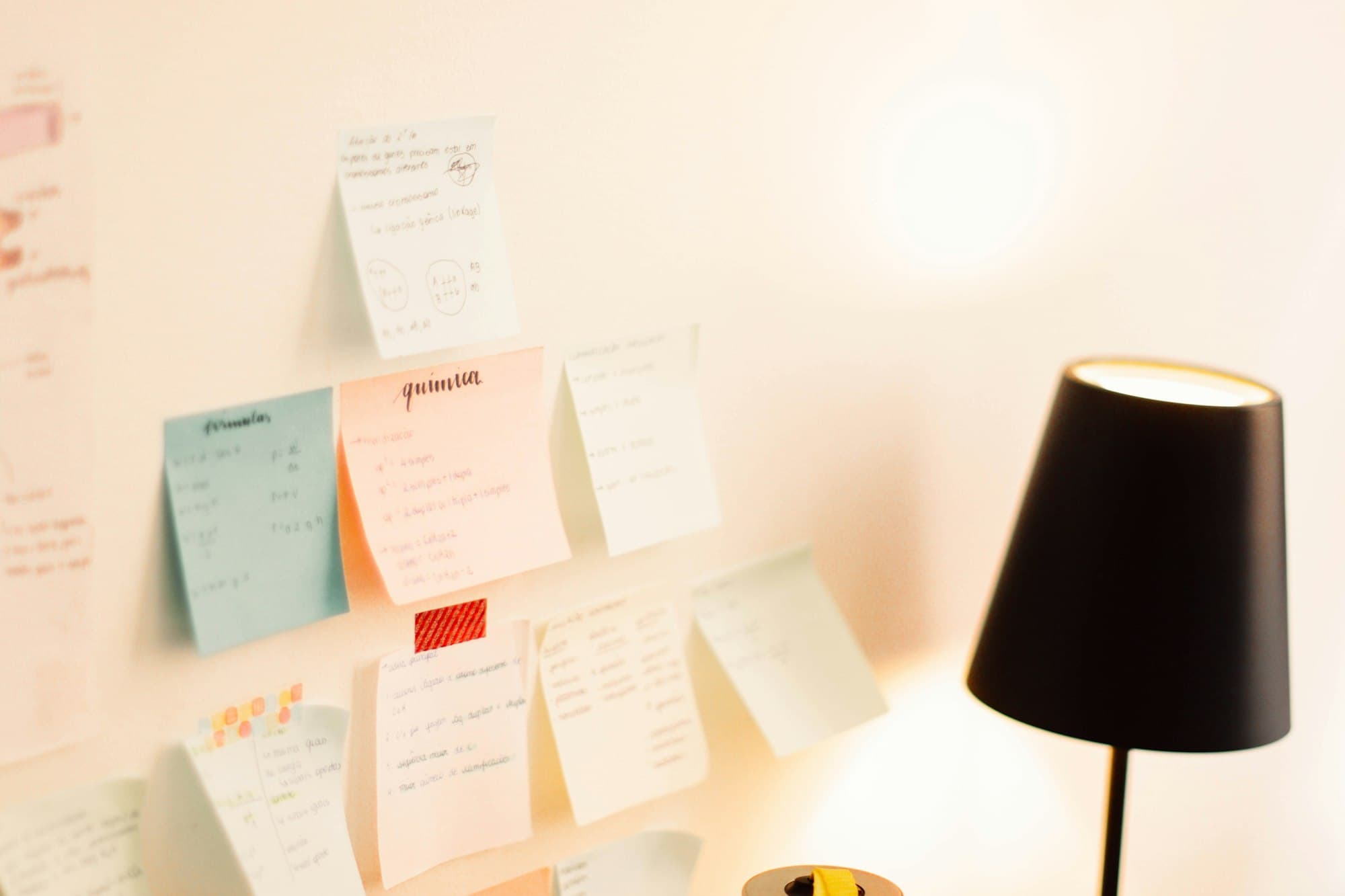 Handwritten sticky notes in pale blue, peach and yellow on a sunlit wall beside a black lamp.