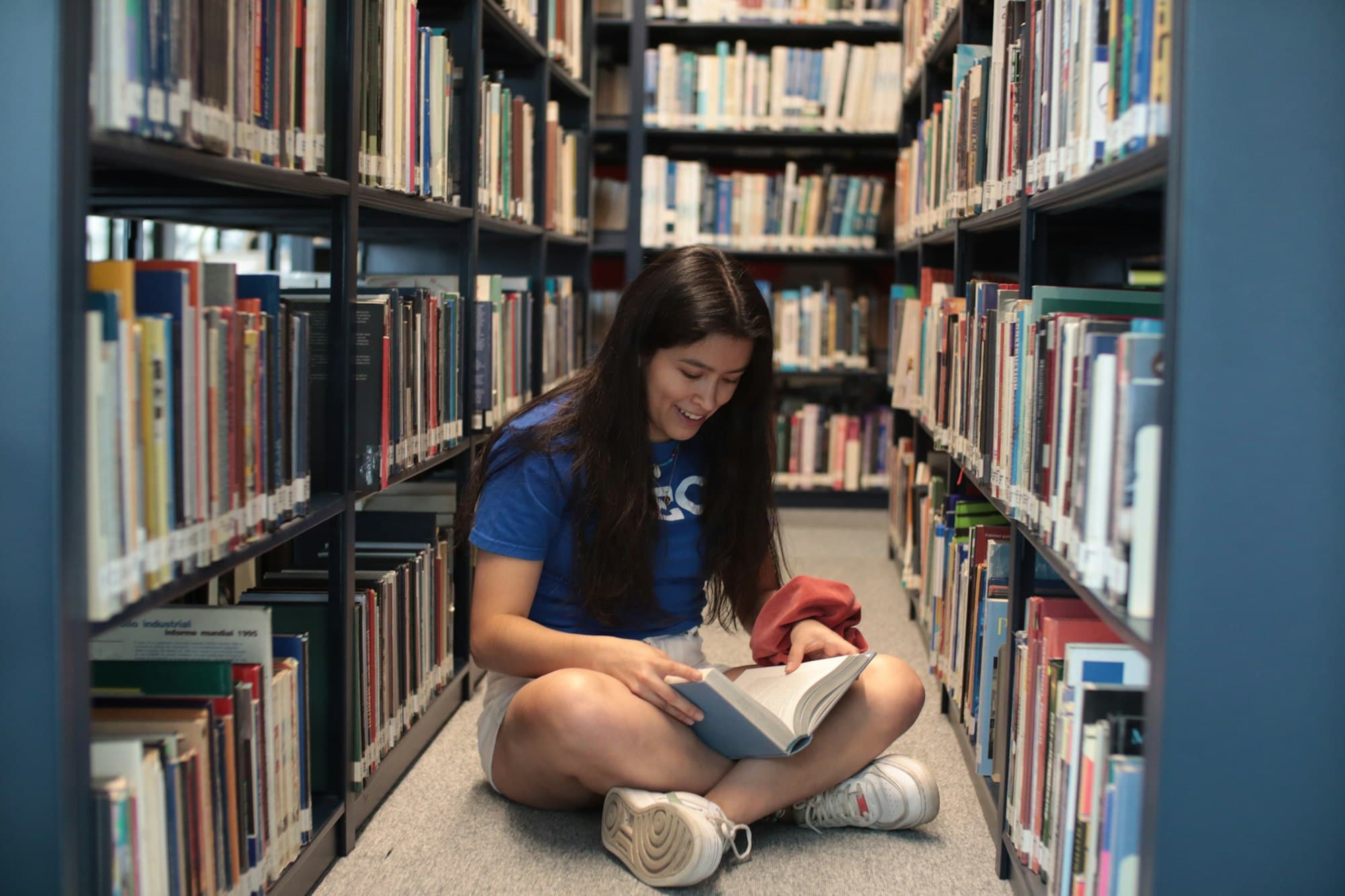 A student sitting cross-legged on a library floor between tall shelves, smiling as she reads a book.